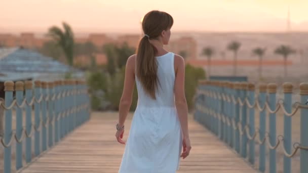 Ung kvinna i vit klänning går en promenad på lång pir vid havet med siluett av bergstoppar på kvällen. Semester och resor koncept. — Stockvideo