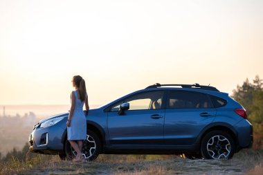 Mavi elbiseli mutlu genç kadın sürücü sıcak yaz akşamının tadını çıkarıyor arabasının yanında duruyor. Seyahat ve tatil kavramı.