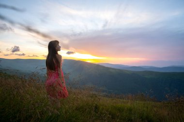 Kırmızılı genç kadın yaz dağlarında rüzgarlı bir akşamda yamaçtaki çayırda yürüyor..