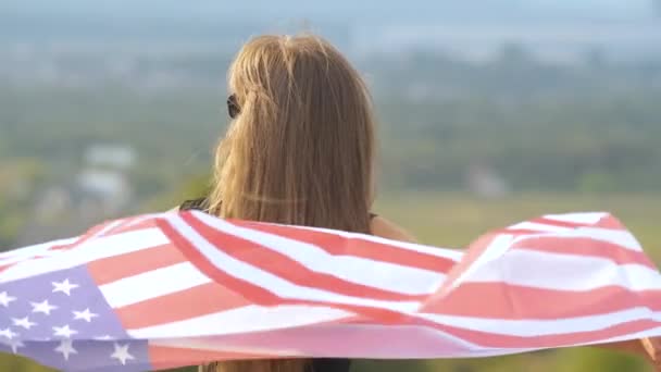 Jovem mulher americana feliz com cabelos longos segurando acenando no vento bandeira nacional dos EUA em seus sholders relaxando ao ar livre desfrutando de dia quente de verão — Vídeo de Stock