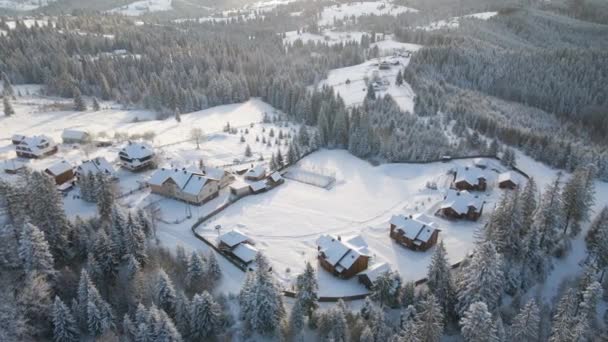Paisaje aéreo de invierno con pequeñas casas de pueblo entre el bosque cubierto de nieve en las montañas frías por la noche — Vídeo de stock