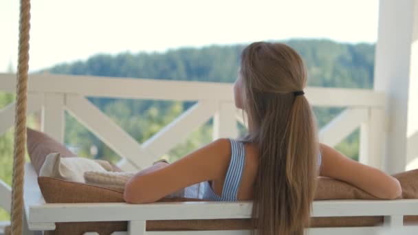 Back view of young brunette woman resting on terrace sofa with soft pillows enjoying view of summer nature. Concept of free time on fresh air — Stock Video