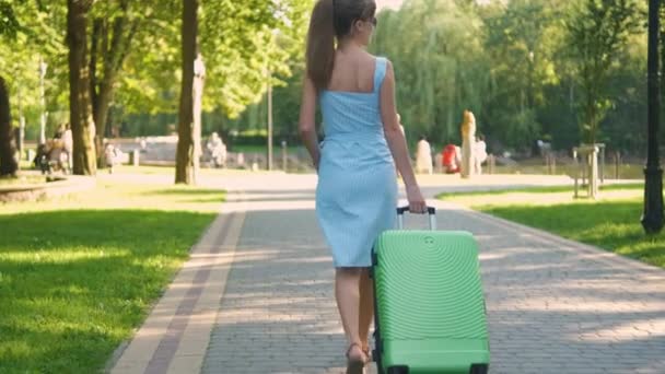 Jeune femme marchant sur le trottoir du parc avec une valise verte le jour de l'été. Concept de voyage et de vacances. — Video