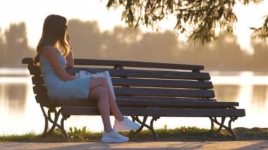 Yaz elbisesi giymiş genç bir kadın sıcak bir akşamda göl kenarında rahat bir şekilde oturuyor. Sağlık, günlük acele kavramından dinleniyor.