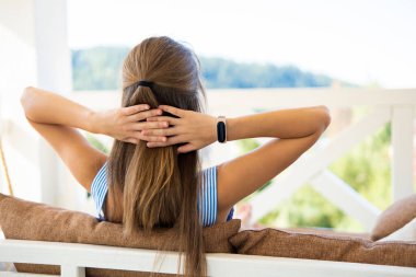 Teras kanepesinde dinlenen esmer bir kadının arkası. Yumuşak yastıkları var. Yaz manzarasının tadını çıkarıyorlar. Temiz havada boş zaman kavramı.