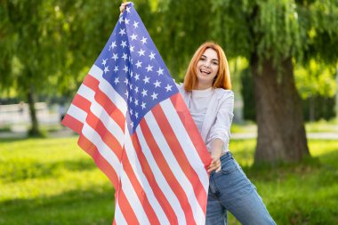 Mutlu kızıl saçlı kadın yaz parkında Amerikan bayrağıyla poz veriyor. ABD 'nin bağımsızlık gününü kutlayan pozitif kız.