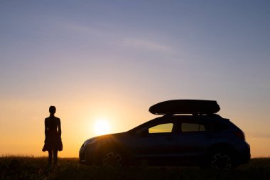 Kadın sürücünün silueti çimenli alanda arabasının yanında duruyor ve gün batımının keyfini çıkarıyor. Genç bayan SUV aracının yanında dinleniyor..