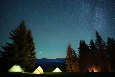 Yürüyüşçülerin silueti, parlak yıldızlı karanlık dağların altındaki kamp alanlarında yanan şenlik ateşinin yanında dinleniyor. Aktif yaşam tarzı ve açık hava yaşam konsepti.
