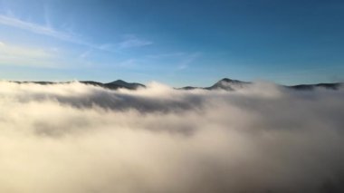 Geniş dağ vadisindeki beyaz kabarık bulutların üzerinde ilerleyen hava görüntüsü.