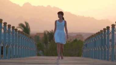 Beyaz elbiseli genç bir kadın uzun rıhtımda yürüyüşe çıkıyor. Akşamları dağ zirveleri siluetiyle. Tatil ve seyahat kavramı.