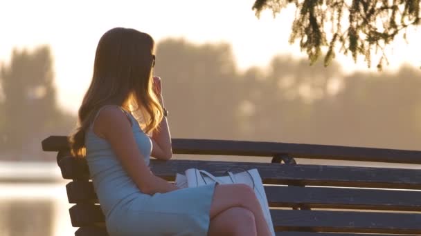 Eenzame vrouw zit alleen op de oever van het meer bank op warme zomeravond. Eenzaamheid en ontspanning in de natuur concept. — Stockvideo