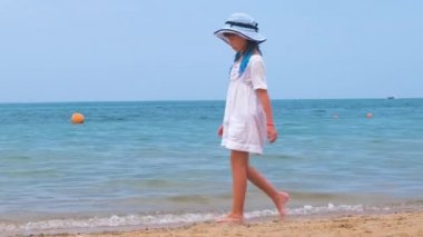 Yaz tatillerinde deniz kıyısında kumsalda yürüyen beyaz elbiseli mutlu bir kız çocuğu. Tatil konseptinde seyahat ve dinlenme