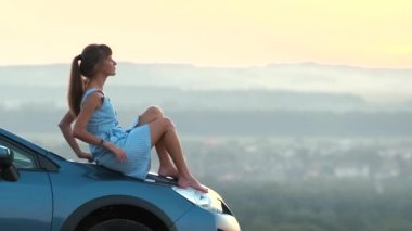 Mavi elbiseli mutlu genç kadın sürücü sıcak yaz akşamının tadını çıkarıyor arabasının kaputunda yatıyor. Seyahat ve tatil kavramı