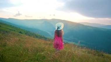 Kırmızı elbiseli mutlu bir kadın yürüyüşçünün arka görüntüsü çimenli bir tepede rüzgarlı bir sonbahar akşamında kollarını açıp güneşin batışının keyfini çıkarırken.