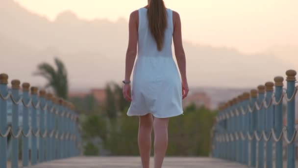 Ung kvinna i vit klänning går en promenad på lång pir vid havet med siluett av bergstoppar på kvällen. Semester och resor koncept — Stockvideo