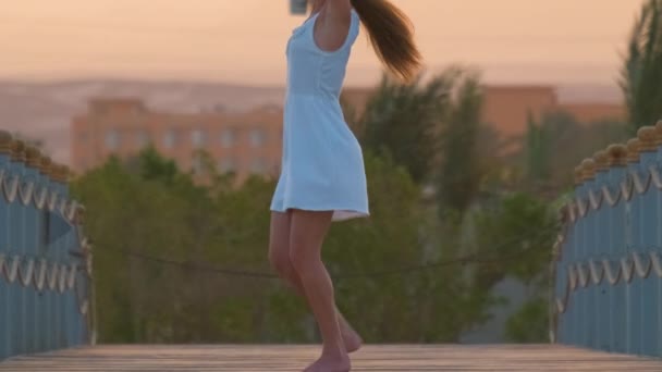 Młoda szczęśliwa kobieta w jasnej sukience tańcząca na długim molo nad morzem z sylwetką górskich szczytów wieczorem. Koncepcja wakacji i podróży — Wideo stockowe