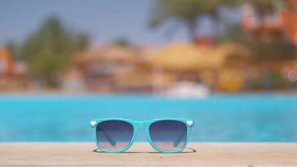 Primer plano de las gafas de sol azules en el lado de la piscina en el complejo tropical en un día soleado cálido. Concepto vacaciones de verano — Vídeo de stock