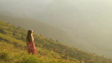 Kırmızı elbiseli genç mutlu kadın gezgin yaz dağlarında rüzgarlı bir akşamda çimenli yamaçta duruyor gün batımında doğanın tadını çıkarıyor.
