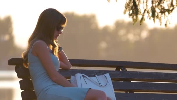 Jonge vrouw in de zomer jurk zitten ontspannen op meer zijbank op warme avond. Wellness rust van het dagelijkse spoedconcept — Stockvideo