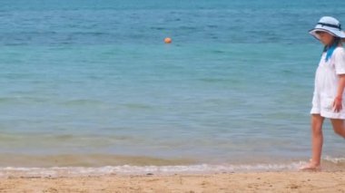 Yaz tatillerinde deniz kıyısında kumsalda yürüyen beyaz elbiseli mutlu bir kız çocuğu. Tatil konseptinde seyahat ve dinlenme