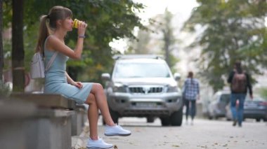 Kağıt bardaktan kahve içen genç bir kadın yaz parkındaki bankta oturuyor.