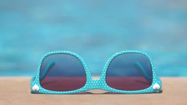 Kék napszemüveg a medence oldalán, meleg napsütéses napon. Nyári szabadság koncepciója — Stock videók