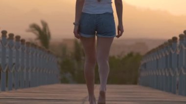 Akşamları deniz kenarında uzun bir rıhtımda yürüyüş yapan ve dağ zirveleri siluetiyle gezen modaya uygun genç bir kadının arka manzarası. Tatil ve seyahat kavramı