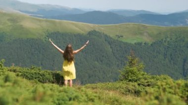 Kırmızılı genç kadın çimenli bir alanda duruyor rüzgarlı bir yaz gününde, dağlarda doğa manzarasının tadını çıkarıyor.