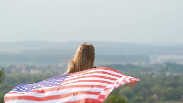 Mladá šťastná žena s dlouhými vlasy drží vlnění na větru americká národní vlajka na její držadla odpočinku venku těší teplý letní den — Stock video