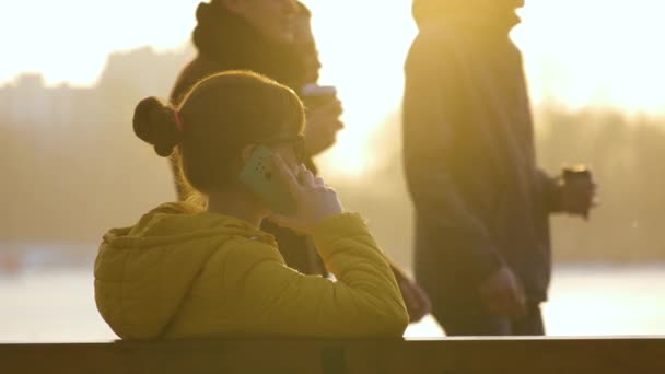 Ung vacker kvinna sitter på en parkbänk och pratar på sin smartphone utomhus under varma höstkvällar — Stockvideo