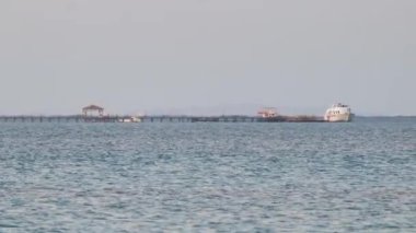 Mavi deniz suyunun dalgalı yüzeyi ve sakin dalgalar üzerinde yüzen uzun rıhtımın sonundaki uzak gemiyle deniz manzarası.