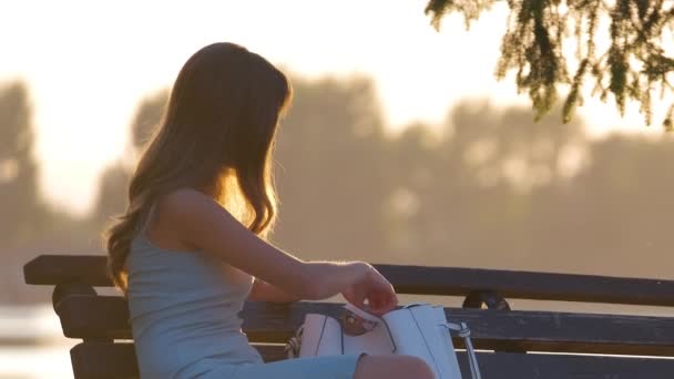 Lonely young woman sitting alone on lake shore bench enjoying warm summer evening. Wellbeing and relaxing in nature concept — Stock Video