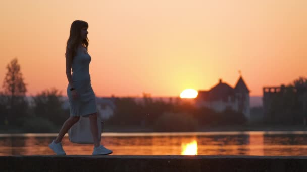Ung kvinna i avslappnad outfit promenader på sjön sida på varm kväll. Sommarsemester och resekoncept — Stockvideo