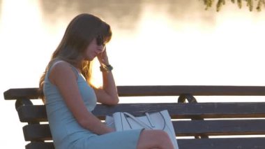 Sıcak bir yaz akşamında, göl kıyısındaki bankta tek başına oturan yalnız üzgün kadın. Yalnızlık ve doğa konseptinde rahatlama.
