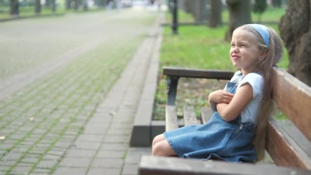 Kis irritált gyermek lány ül egyedül egy padon a nyári parkban — Stock videók