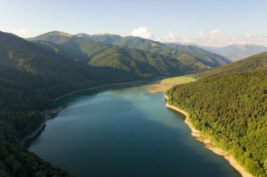 Yoğun yeşil ormanlarla kaplı yüksek dağ tepeleri arasında berrak mavi suyu olan büyük göl manzarası..