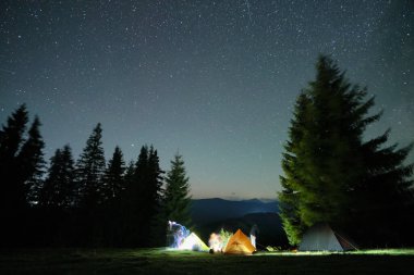 Gece gökyüzünün altındaki karanlık dağ ormanlarında, parlayan yıldızlarla parlayan kamp alanının yakınında parlak turist çadırları. Aktif yaşam tarzı ve açık hava yaşam konsepti.