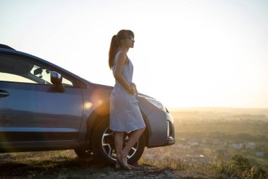 Mavi yazlık elbiseli mutlu kadın sürücü arabasının yanında sıcak bir akşamın tadını çıkarıyor. Seyahat ve tatil kavramı.