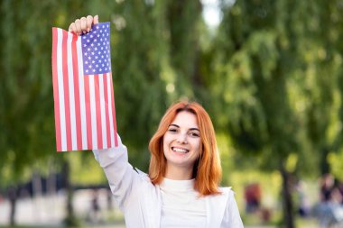 Amerikan bayrağıyla poz veren mutlu genç bir kadın onu açık havada, yaz parkında tutuyor. ABD 'nin bağımsızlık gününü kutlayan pozitif kız.