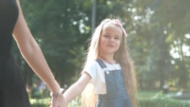 Anne ve uzun saçlı küçük kızı yaz parkında el ele yürüyorlar.