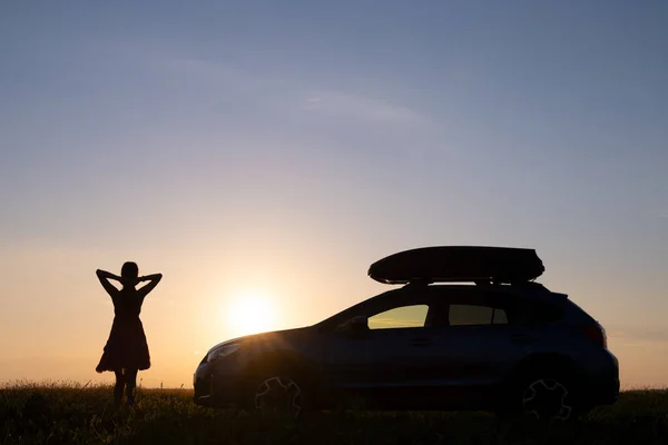 Silueta oscura de mujer conductora de pie cerca de su coche en el campo de hierba disfrutando de la vista de la puesta de sol brillante. Mujer joven relajándose durante el viaje por carretera junto al vehículo SUV — Foto de Stock