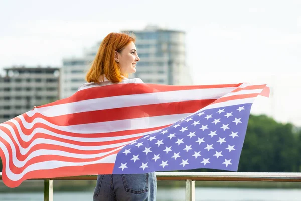 Szczęśliwa rudowłosa dziewczyna z flagą USA na ramionach. Pozytywna młoda kobieta świętująca dzień niepodległości Stanów Zjednoczonych. Międzynarodowy Dzień Koncepcji Demokracji — Zdjęcie stockowe