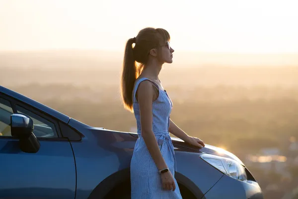 Conductora joven feliz en vestido azul disfrutando de la cálida noche de verano de pie junto a su coche. Concepto de viaje y vacaciones — Foto de Stock