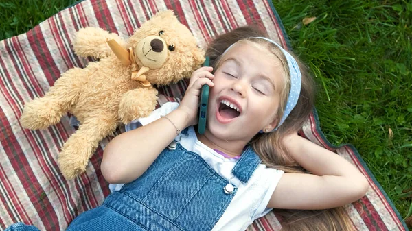 작고 행복 한 여자 아이가 잔디에 누워 테디 베어 와 휴대폰으로 얘기하는 모습 — 스톡 사진