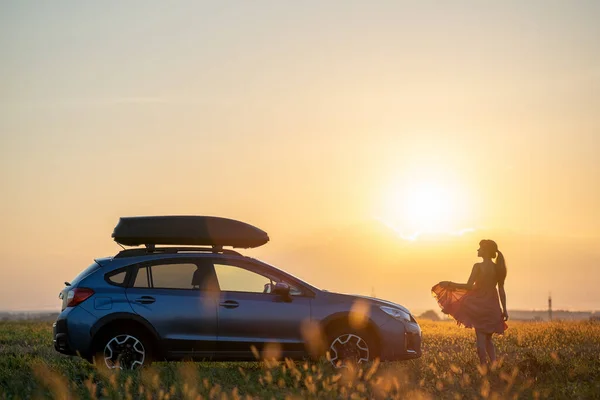 Silueta de conductora de pie cerca de su coche en el campo de hierba disfrutando de la vista de la puesta de sol brillante. Mujer joven que se relaja durante el viaje por carretera junto al vehículo SUV — Foto de Stock