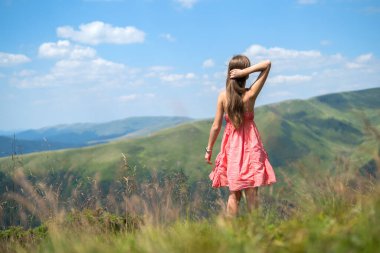 Yaz dağlarında rüzgarlı bir günde çimen tarlasında yürüyen kırmızı elbiseli mutlu genç bir kadın.