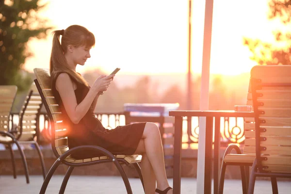 Молодая женщина, сидящая за столом уличного кафе, просматривает свой мобильный телефон под открытым небом теплым летним вечером. Концепция связи и мобильной связи — стоковое фото