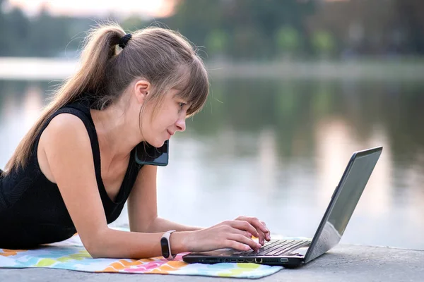 Mujer joven que trabaja detrás de la computadora portátil mientras habla en el teléfono móvil acostado en el parque de verano al aire libre — Foto de Stock