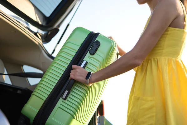 Junge Frau im Sommerkleid steckt grünen Koffer in den Kofferraum ihres Autos. Reise- und Urlaubskonzept — Stockfoto