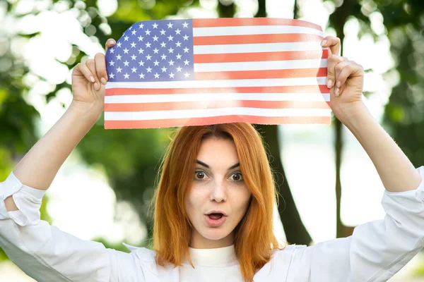 Wściekła młoda ruda kobieta protestująca z flagą USA nad głową stojąca na świeżym powietrzu w letnim parku. Aktywistka na demonstracji z chorągwią narodową — Zdjęcie stockowe